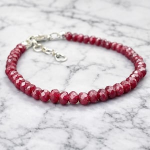 Puede incluir: Una pulsera de cuentas de color rojo rubí con un cierre plateado. Las cuentas facetadas son de color rojo intenso y están ensartadas para formar una delicada joya. La pulsera se muestra sobre una superficie de mármol.