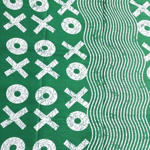 Op de afbeelding: Een levendige groene stof met een herhalend wit patroon van X- en O-symbolen met een gebarsten textuureffect. Aan de rechterkant toont een deel van het textiel een reeks verticale, golvende witte lijnen die contrasteren met de geometrische vormen.