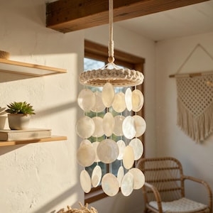 May include: A decorative wind chime featuring a circular woven top and multiple strands of white, oval-shaped shell pieces. The chime hangs from a natural rope, creating a coastal or bohemian style. The shells have an iridescent sheen.