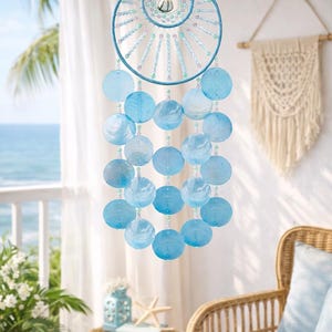 Puede incluir: Un carillón de viento colgante de temática costera. El carillón presenta un marco circular azul con un círculo interior más pequeño, adornado con cuentas y conchas redondas azules translúcidas colgantes. El diseño evoca una estética playera y náutica.