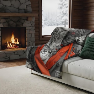 Op de afbeelding: Een decoratieve sprei met een gedetailleerde illustratie van een vrouw met tatoeages, een rode sjerp en een wolf. De sprei is gedrapeerd over een witte bank, met een groen kussen. De achtergrond omvat een open haard en een besneeuwd raam.