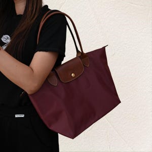 May include: A burgundy tote bag with brown leather detailing. The bag has a flap closure with a gold button and brown leather handles. The bag is made of a smooth, durable material, and is designed for everyday use.