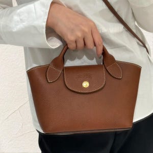 May include: A brown leather handbag with a flap closure and a gold-coloured button. The bag has two handles and a shoulder strap. The stitching is white, and the bag is a medium size.