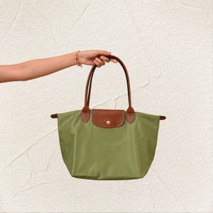 May include: Olive green tote bag with brown leather detailing on the flap and handles. The bag has a rectangular shape with a fold-over flap secured by a gold-coloured button. The handles are long enough to be worn over the shoulder.