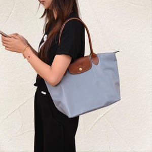 May include: A light grey tote bag with brown leather detailing. The bag features a flap closure and brown leather handles. The bag is made from a durable material, suitable for daily use.