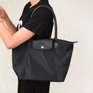 May include: Black tote bag with leather detailing. The bag has a flap closure with a silver button and a top handle. The bag is made of a nylon material and has a simple, elegant design.