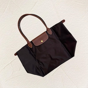 May include: A black tote bag with brown leather handles and a flap closure. The bag has a brown leather flap with a gold-coloured snap closure. The handles are curved and attached to the top of the bag.