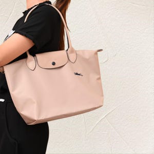 May include: A blush-coloured tote bag with a flap closure and black accents. The bag features a black logo on the front and has two handles. The bag is made of a smooth material and has a rectangular shape.