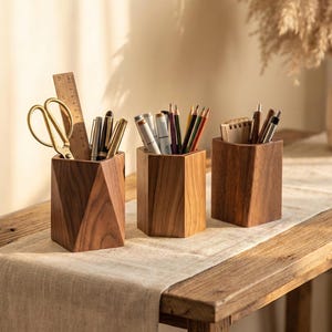 Peut inclure: Trois organiseurs de bureau en bois contenant diverses fournitures de bureau. Les organisateurs sont de forme géométrique et en bois foncé. Le premier contient des ciseaux dorés, une règle et des stylos. Le second contient des stylos et des crayons. Le troisième contient des stylos et un petit bloc-notes.