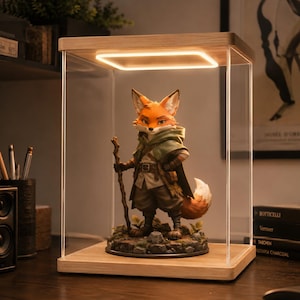 May include: A fox figurine standing on a rocky base inside a clear acrylic display case with a light wood base and top. The fox wears a green hooded cloak, a brown tunic, and holds a wooden staff. A square LED light is integrated into the top.