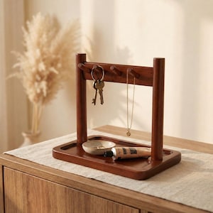 Peut inclure: Un organiseur de clés et de bijoux en bois brun foncé, avec une base rectangulaire et des supports verticaux. Des clés et un collier sont suspendus en haut. Un petit plat et un tube de crème sont posés sur la base.