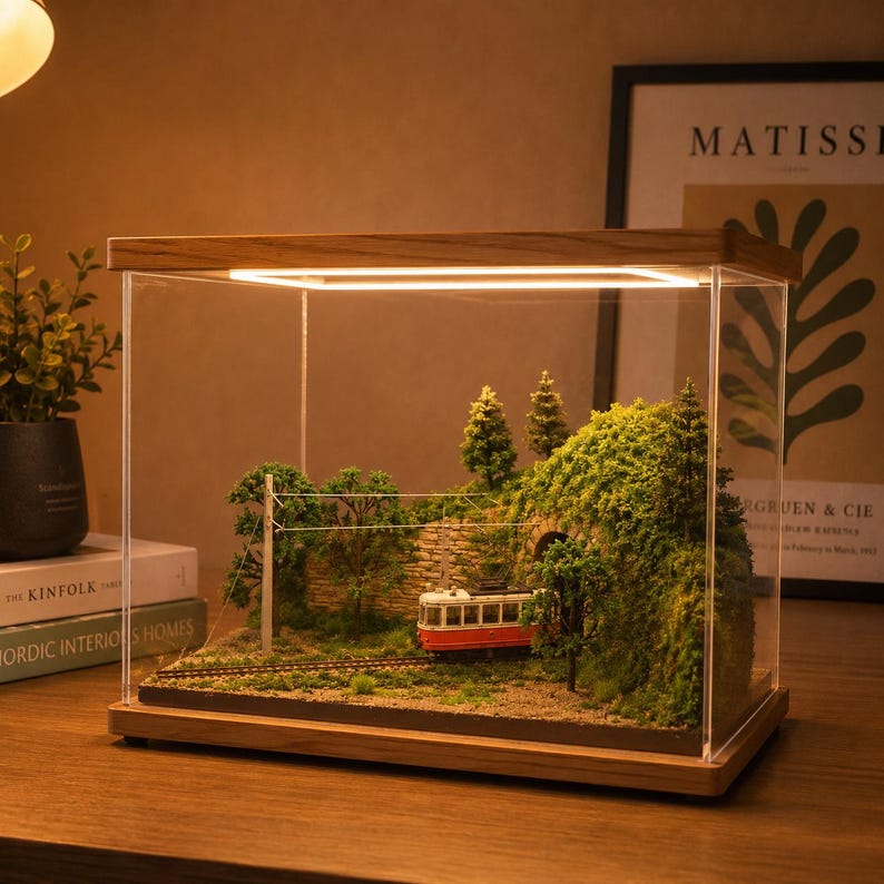 May include: A miniature diorama encased in a clear acrylic box with a wooden base and top, featuring a red and white tram on tracks emerging from a stone tunnel entrance surrounded by green trees and foliage, illuminated by an internal light strip.