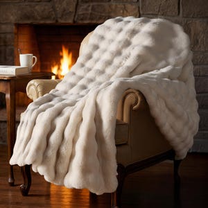 May include: A cream-colored, plush throw blanket draped over a leather armchair. The blanket has a textured, quilted pattern and a soft, furry appearance. A fireplace with a burning fire is in the background, creating a cozy atmosphere.