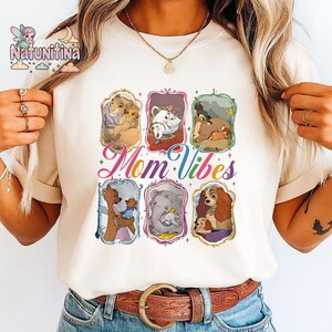 Può includere: T-shirt color crema con sei illustrazioni incorniciate di personaggi animati con i loro figli. La frase "Mom Vibes" è scritta in uno script colorato sotto le illustrazioni. La maglietta ha un girocollo e maniche corte.