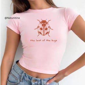 May include: A light pink t-shirt featuring a brown ladybug graphic with white spots. The text "the last of the bugs" is printed below the ladybug. The shirt has short sleeves and a crew neck.