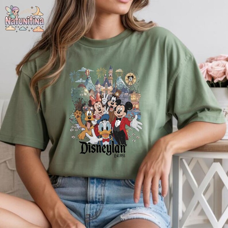 May include: Sage green t-shirt featuring a colorful graphic of Disney characters including Mickey Mouse, Minnie Mouse, Donald Duck, and Goofy. The graphic includes the Disneyland logo and various park attractions. The shirt is a relaxed fit.