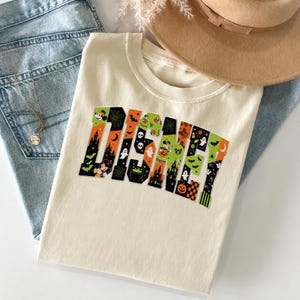 Pode incluir: Uma t-shirt cor de creme com a palavra DISNEY em letras grandes de bloco. As letras estão preenchidas com um padrão temático de Halloween, incluindo fantasmas, abóboras, morcegos, teias de aranha, castelos e ícones do Mickey Mouse.
