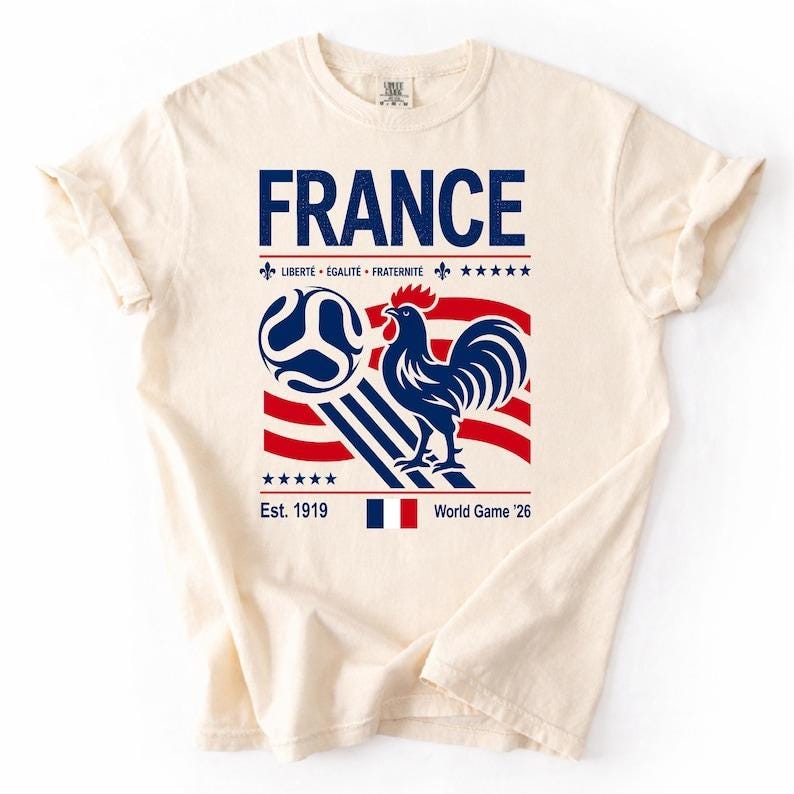 May include: A cream-colored t-shirt featuring a graphic with the word FRANCE in blue, a blue rooster, a soccer ball, and red and blue stripes. The text includes LIBERT&Eacute; &Eacute;GALIT&Eacute; FRATERNIT&Eacute;, Est. 1919, and World Game '26, along with a small French flag.