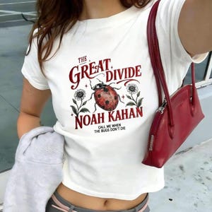Op de afbeelding: Een wit T-shirt met korte mouwen en een rood-zwarte afbeelding van een lieveheersbeestje geflankeerd door twee bloemen. De tekst luidt THE GREAT DIVIDE, NOAH KAHAN en CALL ME WHEN THE BUGS DON'T DIE. Een rode schoudertas wordt gedragen.