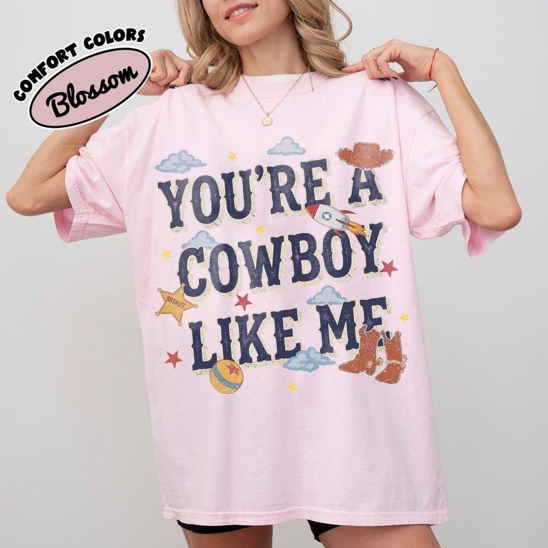 May include: A light pink short-sleeve t-shirt featuring the text "YOU'RE A COWBOY LIKE ME" in dark blue letters. The graphic includes illustrations of a cowboy hat, a rocket, a sheriff star, a ball, and cowboy boots, surrounded by small stars and clouds.