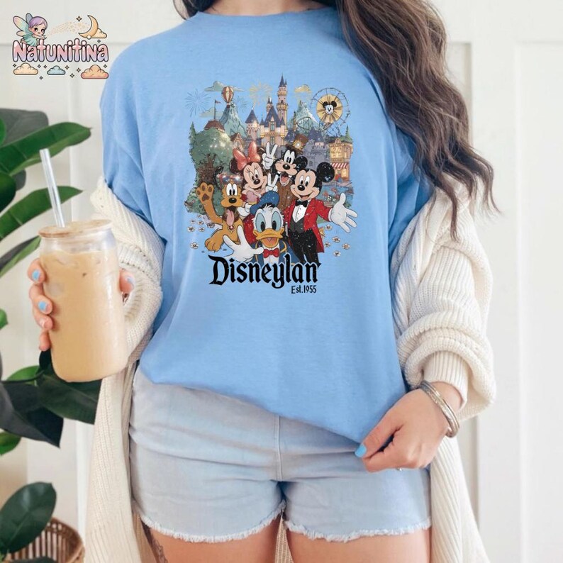 May include: Light blue t-shirt featuring a colorful graphic of Disney characters in front of a castle and amusement park. The word "Disneyland" is printed in a stylized font, with "Est. 1955" below.