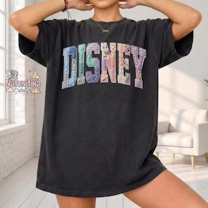 May include: A black oversized t-shirt featuring the word "DISNEY" in a patchwork design. The letters are in pastel colours and patterns. The shirt has short sleeves and a relaxed fit, suitable for casual wear.