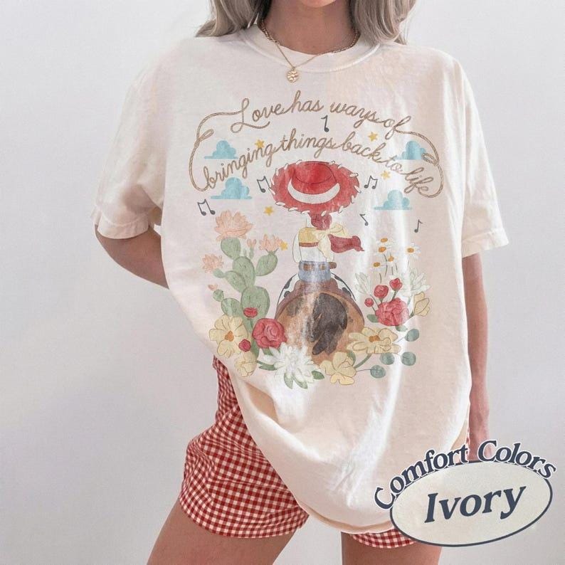 May include: An ivory t-shirt featuring a graphic print of a cowgirl character in a red hat, surrounded by cacti and flowers. The text reads, Love has ways of bringing things back to life, accompanied by musical notes and small blue clouds.