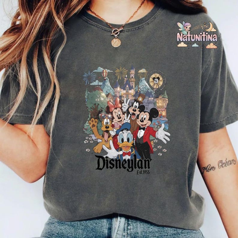 May include: Dark gray t-shirt featuring a colorful graphic of Disney characters including Mickey Mouse, Minnie Mouse, Donald Duck, and Goofy in front of a castle and fireworks. The text "Disneyland Est. 1955" is below the graphic.