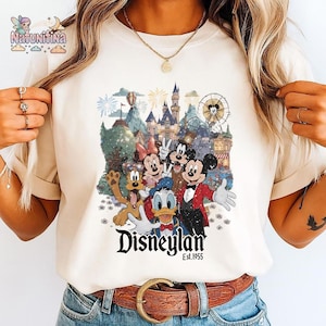 May include: Cream-colored t-shirt featuring a colorful graphic of Disney characters, including Mickey Mouse, Minnie Mouse, Donald Duck, and Goofy, in front of a castle and fireworks display. The word "Disneyland" is printed in a stylized font, with "Est.1955" below.