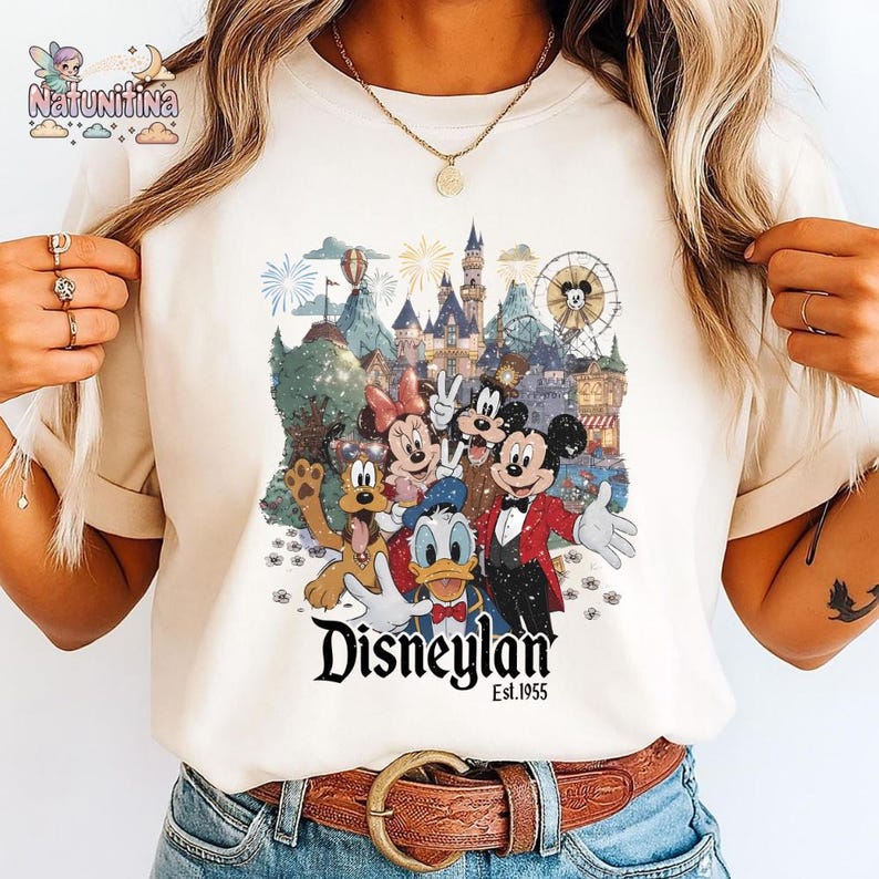 May include: Cream-colored t-shirt featuring a colorful graphic of Disney characters, including Mickey Mouse, Minnie Mouse, Donald Duck, and Goofy, in front of a castle and fireworks display. The word "Disneyland" is printed in a stylized font, with "Est.1955" below.