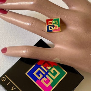 May include: A gold-coloured ring featuring a multi-coloured four-square logo. The logo is red, green, blue, and yellow. The ring is on a hand with red nail polish. The ring is in a black box with the word "GIVENCHY" printed on it.