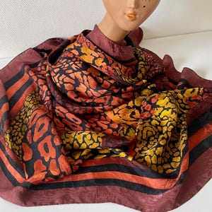 Peut inclure: Un foulard carré en soie avec un imprimé animalier audacieux dans les tons orange, jaune et noir. Le foulard a une bordure bordeaux avec des rayures noires et orange. Le foulard est drapé sur une tête de mannequin.