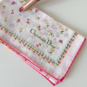 May include: White handkerchief with a floral pattern of pink and yellow flowers, and a pink border. The text "Christian Dior" is printed in green. The handkerchief is folded and displayed on a white surface.