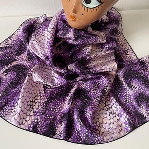 Könnte beinhalten: Ein quadratisches Tuch mit einem sich wiederholenden Schlangenhautmuster in Violett, Schwarz und Hellrosa mit einem dünnen schwarzen Rand. Das Tuch ist um den Hals eines stilisierten Schaufensterpuppenkopfes mit braunem Haar, großen Augen und orangefarbenen Lippen drapiert.