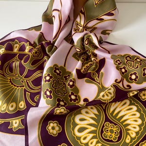 Puede incluir: Un pañuelo de seda vintage con un estampado floral en tonos lavanda, verde oliva, dorado y morado oscuro. El pañuelo está drapeado sobre una cabeza de maniquí, mostrando su diseño y textura. Los bordes del pañuelo están acabados con flecos.