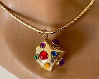 Puede incluir: Collar dorado con un colgante geométrico adornado con piedras preciosas de colores: rojo, verde, azul, amarillo y morado. El collar se muestra sobre un maniquí.