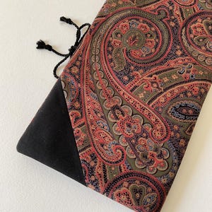 Może przedstawiać: Materiałowe etui z wzorem paisley w odcieniach czerwieni, zieleni i niebieskiego. Etui ma czarną, trójkątną podstawę i zamknięcie na sznurek. Wzór paisley jest szczegółowy i pokrywa większość powierzchni.