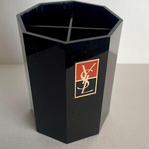 May include: A black octagonal desk organiser with an internal cross-shaped divider. The front features a rectangular emblem with a red top half and black bottom half, displaying the gold YSL logo and a thin gold border around the entire emblem.