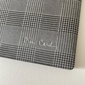 May include: Close-up of a fabric with a black and white checkered pattern. The fabric has a houndstooth design with the words "Pierre Cardin" in cursive script. The fabric is likely used for clothing or accessories.