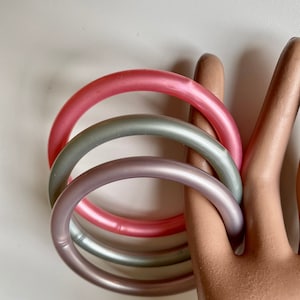 Peut inclure: Trois bracelets de couleur pastel, rose, argent et lavande, sont entrelacés. Les bracelets ronds et lisses sont présentés sur un fond neutre. Les bracelets ont un diamètre d'environ 7,6 cm.