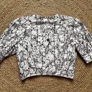 May include: A white blouse with a black floral pattern. The blouse has three-quarter sleeves with tie details at the cuffs. The fabric is lightweight and the blouse is laid flat on a textured surface.