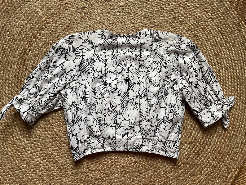 May include: A white blouse with a black floral pattern. The blouse has three-quarter sleeves with tie details at the cuffs. The fabric is lightweight and the blouse is laid flat on a textured surface.