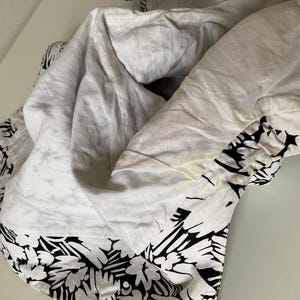 May include: White fabric with a black floral pattern along the edge. The fabric is folded and appears to be a piece of clothing or bedding. The white fabric has some discoloration.