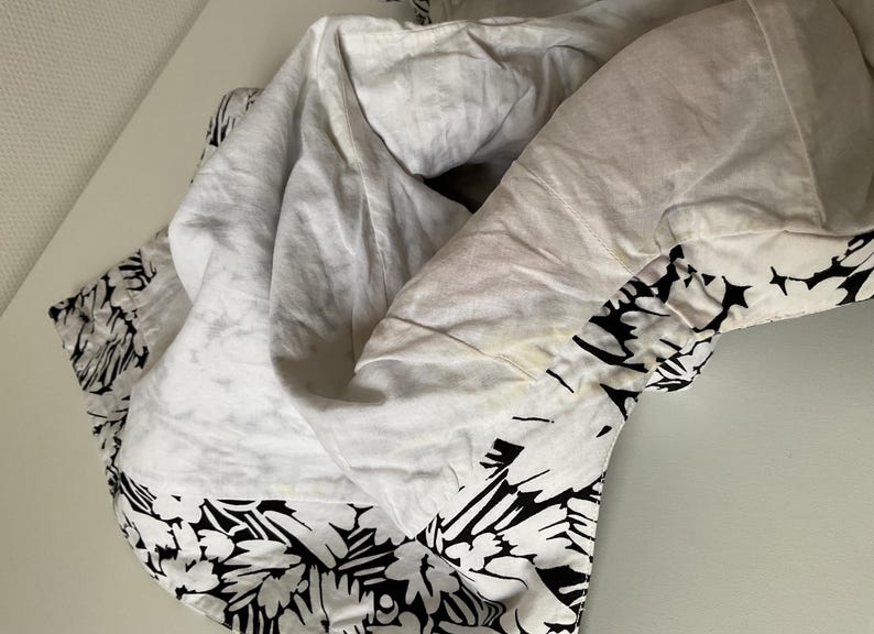 May include: White fabric with a black floral pattern along the edge. The fabric is folded and appears to be a piece of clothing or bedding. The white fabric has some discoloration.