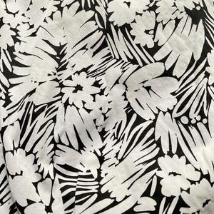 May include: Close-up of a fabric with a bold black and white floral pattern. The design features large, stylized flowers and leaves against a white background. The fabric appears to be soft and possibly cotton or linen.