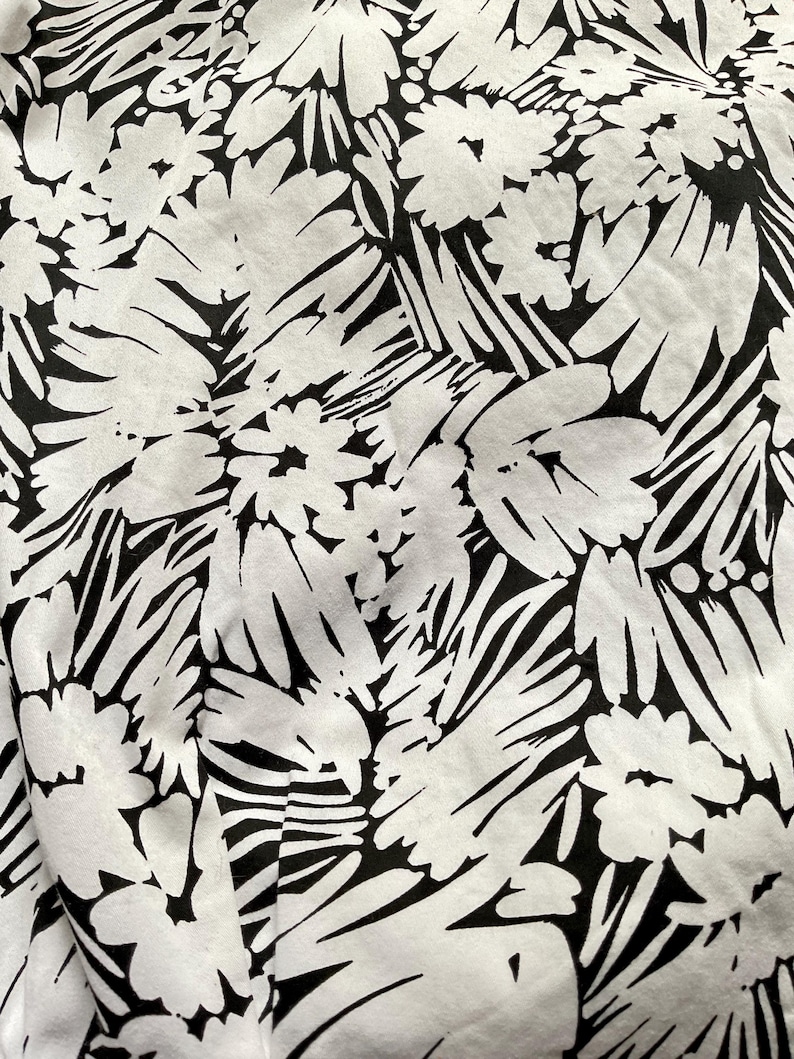May include: Close-up of a fabric with a bold black and white floral pattern. The design features large, stylized flowers and leaves against a white background. The fabric appears to be soft and possibly cotton or linen.