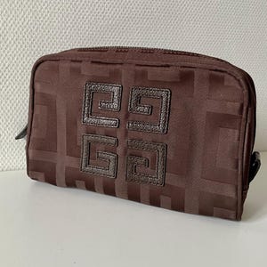 May include: A brown rectangular cosmetic bag with a zip closure. The bag features a repeating geometric pattern and a dark brown leather emblem on the front. The emblem is made up of four connected squares. The bag is made of a textured fabric.