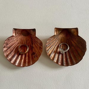 Könnte beinhalten: Zwei kupferfarbene, muschelförmige Ringteller. Ein Teller hat einen goldenen Ring drin, der andere einen silbernen Ring mit einem klaren Stein.
