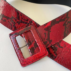 Peut inclure: Une ceinture en cuir de serpent rouge avec une grande boucle rectangulaire. La ceinture est fabriquée en France par Atelier Bower. Le texte "I 1 70H 08" est imprimé au dos de la ceinture.