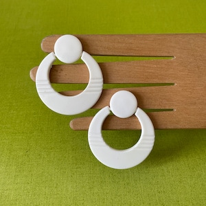 Può includere: Un paio di orecchini a cerchio in plastica bianca con un design circolare. Gli orecchini hanno una finitura liscia e opaca.